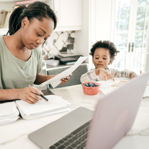 A women doing paperwork in front a laptop while a baby sits at the table with a snack